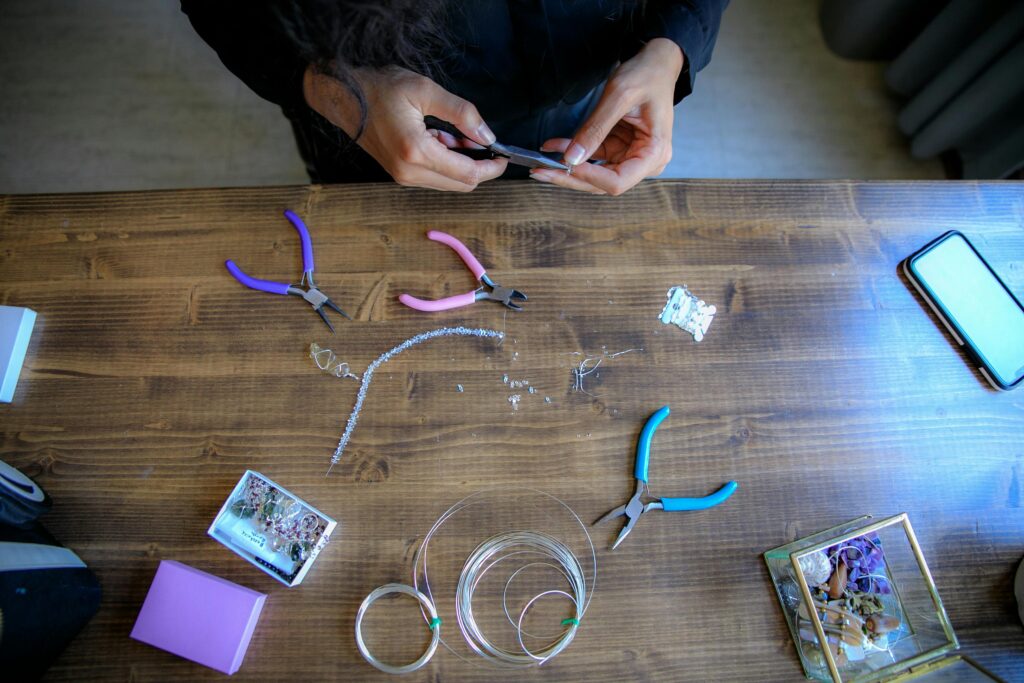 A woman crafting jewelry using tools at a wooden desk, viewed from above.
