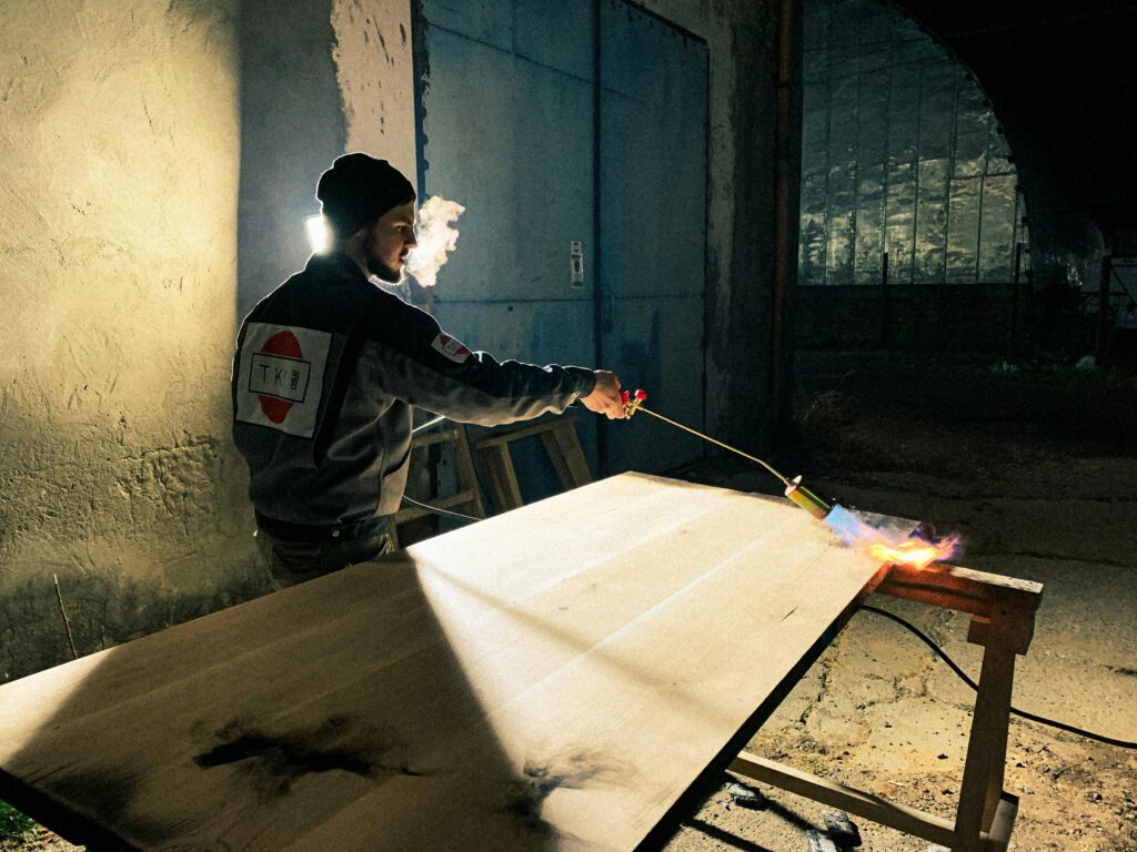 A craftsman uses a blowtorch to treat wood indoors, creating a dramatic effect.