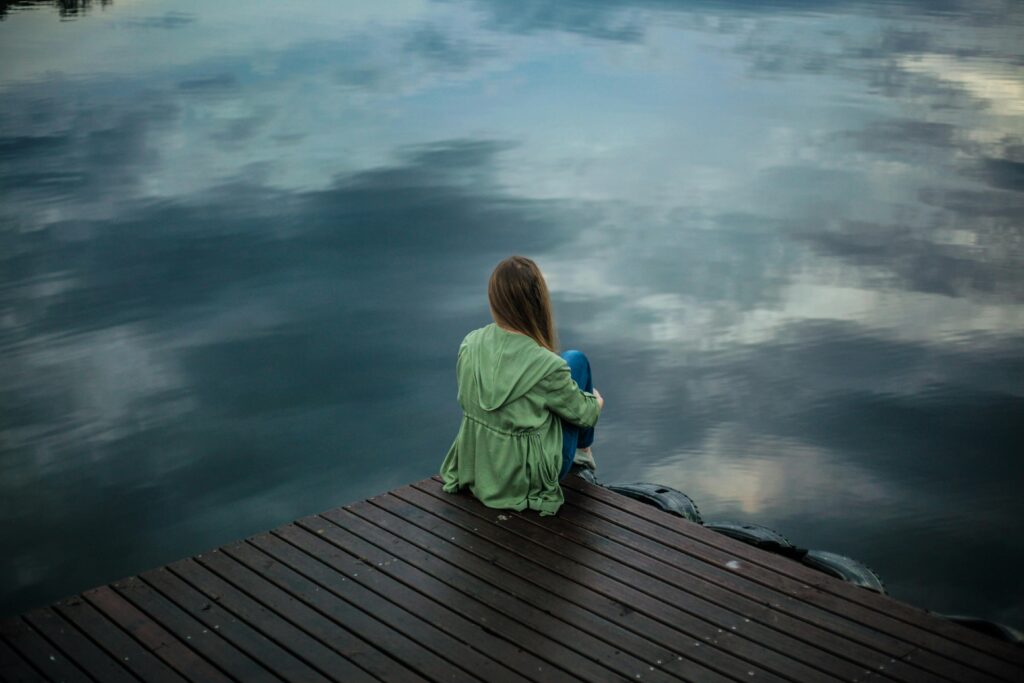 Natural Relief for Stress and Anxiety: Popular Supplements and Devices A woman sits on a wooden dock, reflecting by a calm lake under a cloudy sky.